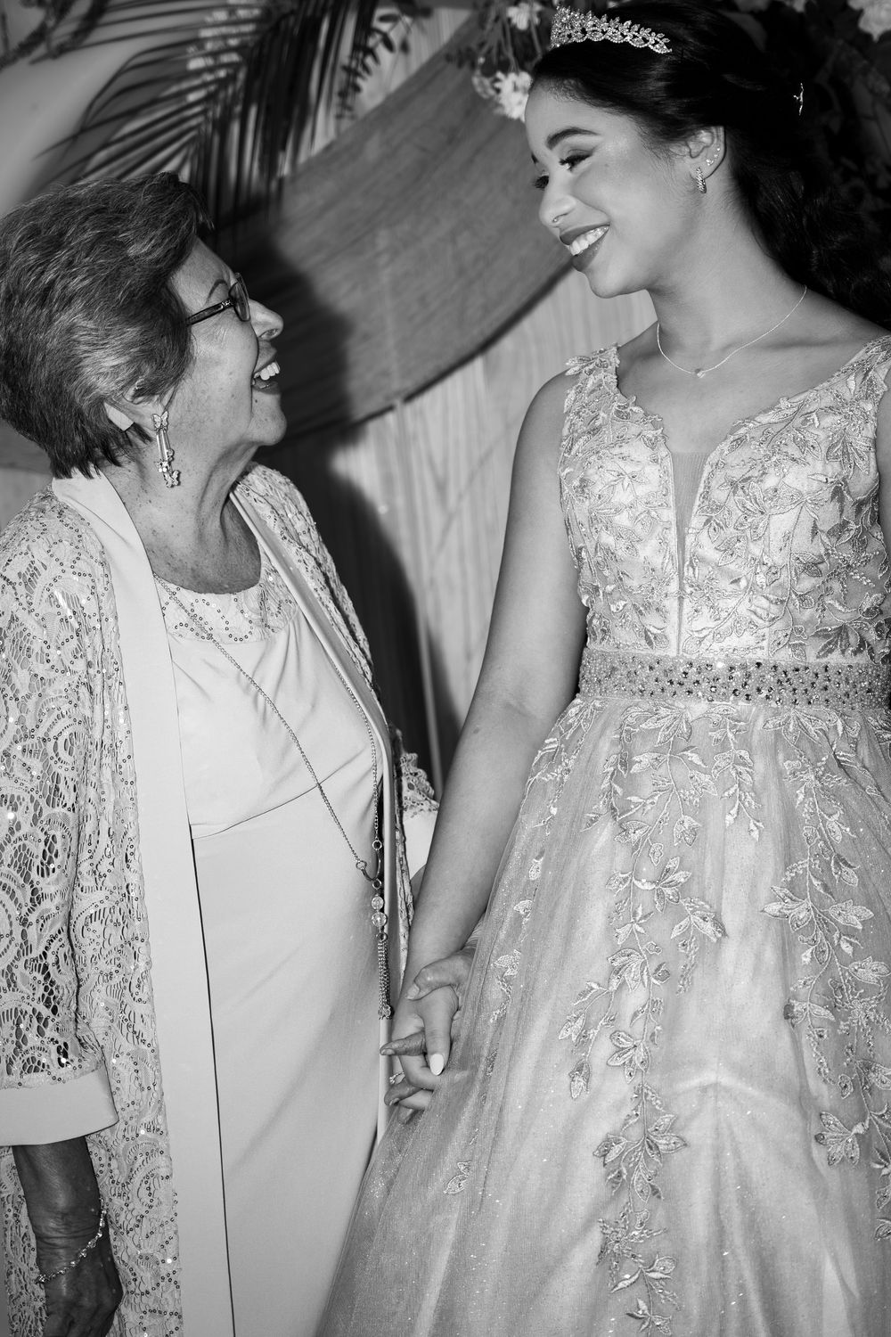 Black and white photo of a bride in a tiara and lace gown smiling and holding hands with an elderly woman.