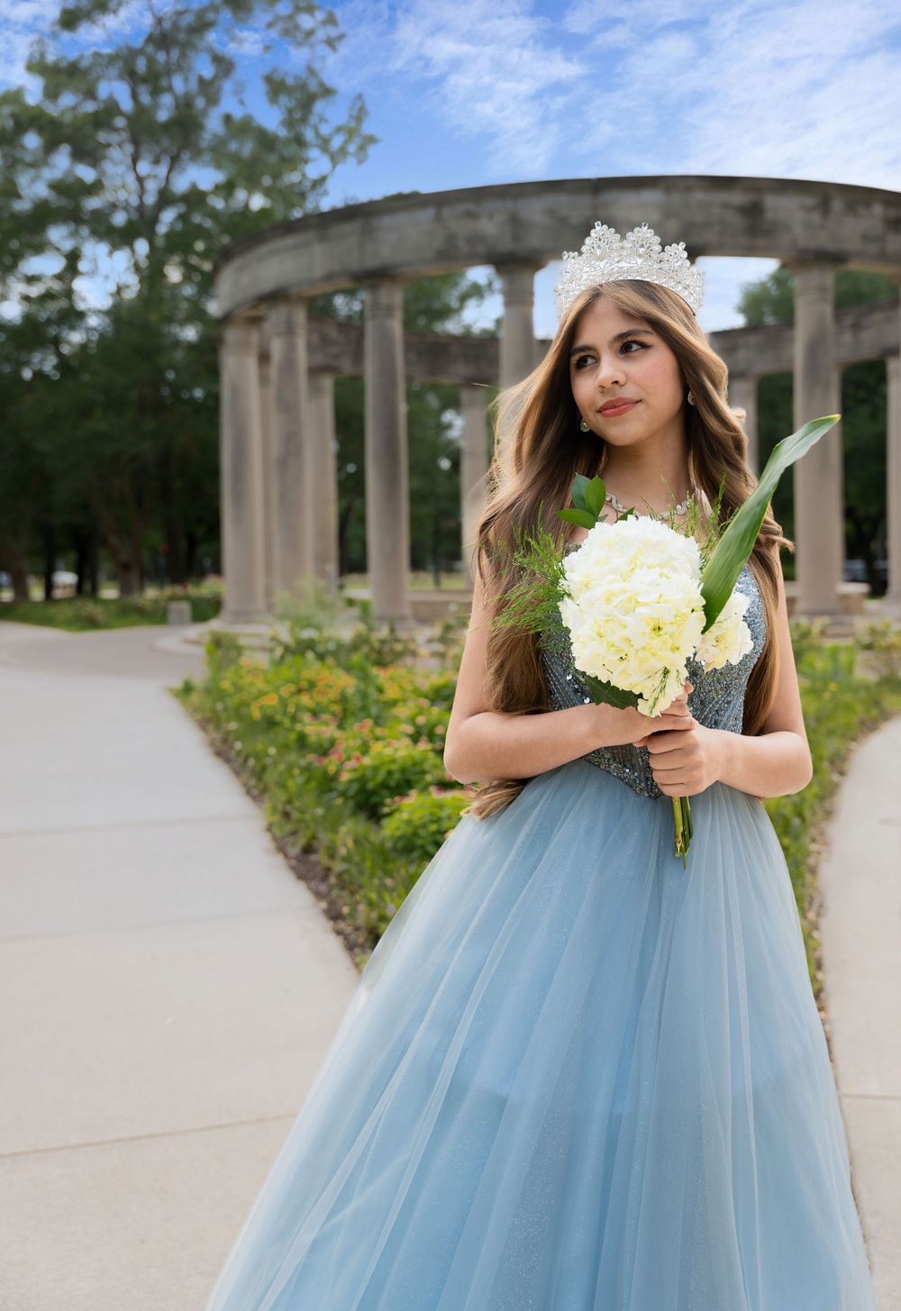 Young woman in blue tulle gown and crystal crown holds white bouquet near stone columns outdoors.