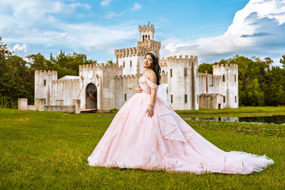 Young woman in pink ball gown poses before a white castle on lush green grounds under a blue sky.