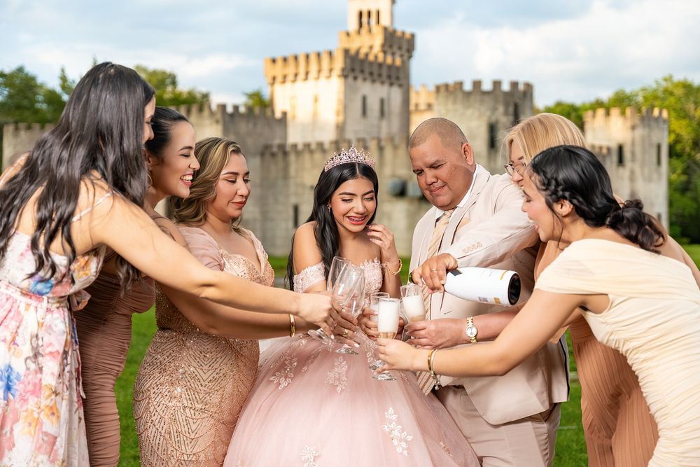 Quinceañera girl in pink gown celebrates with family and friends toasting champagne in front of a castle.