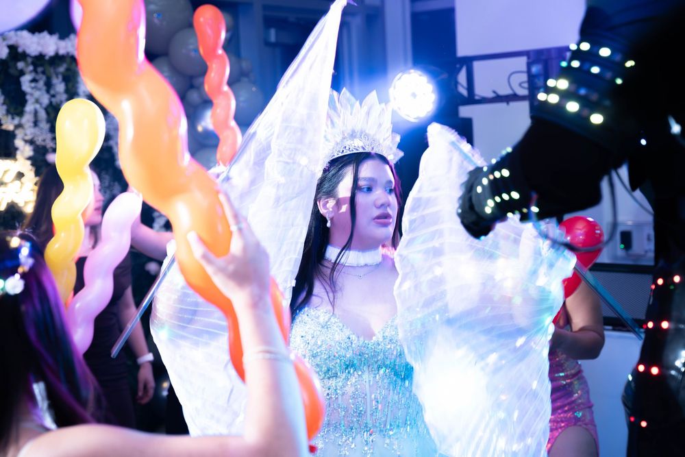 A woman in a glowing white angel costume with light-up wings performs at a colorful nighttime carnival event.