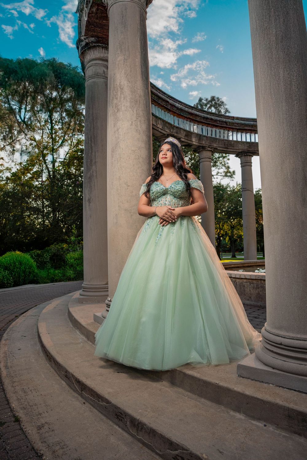 Young woman in mint green quinceañera ball gown poses between stone columns in an elegant outdoor setting.