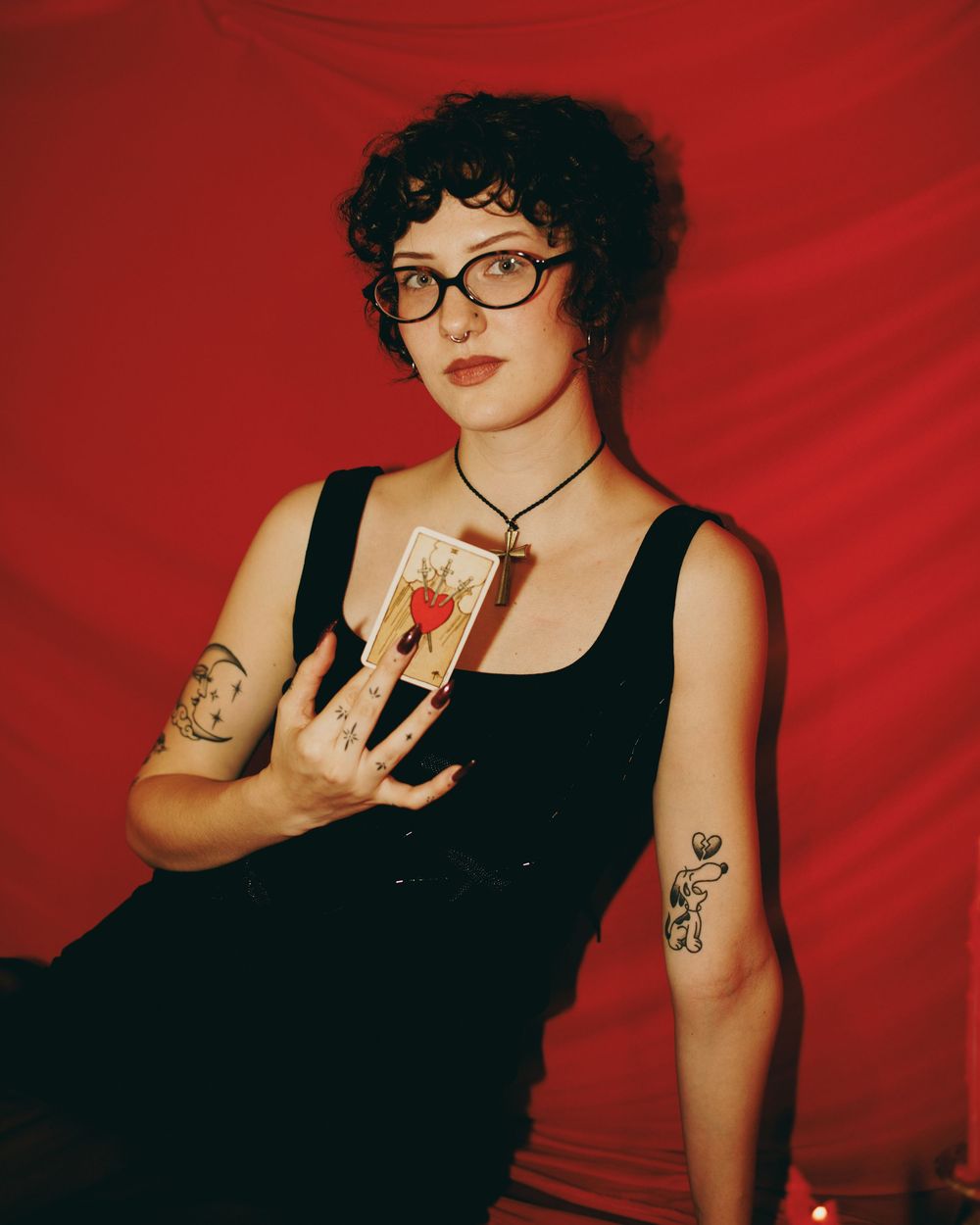 Woman with curly hair, glasses, and tattoos holds a tarot card against a red background.