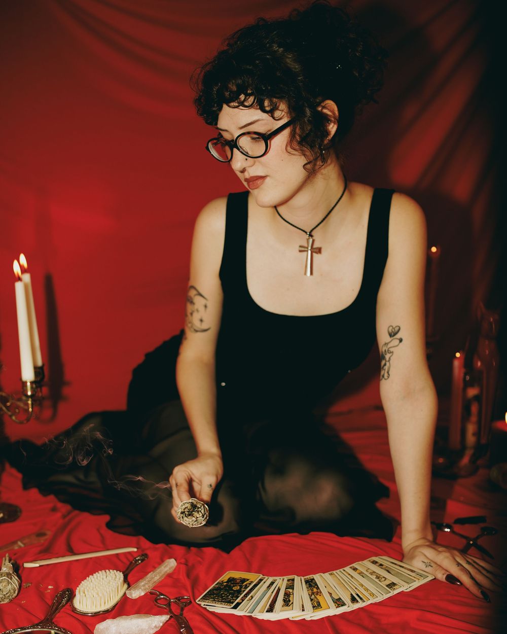 Woman with glasses and cross necklace reads tarot cards on red fabric surrounded by candles and mystical objects.