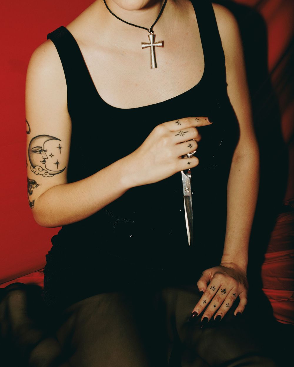 Woman in black tank top with cross necklace, tattoos, and rings holding a dagger against red background.