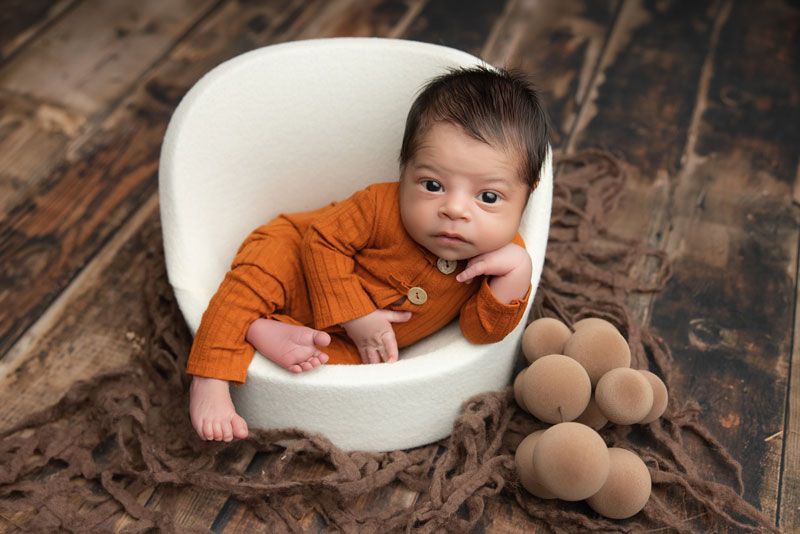 Newborn baby in orange outfit posed in white bowl prop on brown wood floor with decorative eggs.