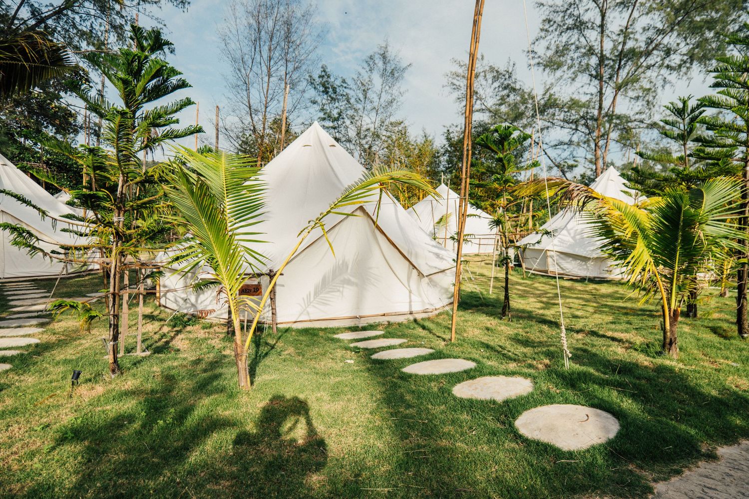 White bell tents set among tall palm trees on lush green grass at a tropical glamping resort.