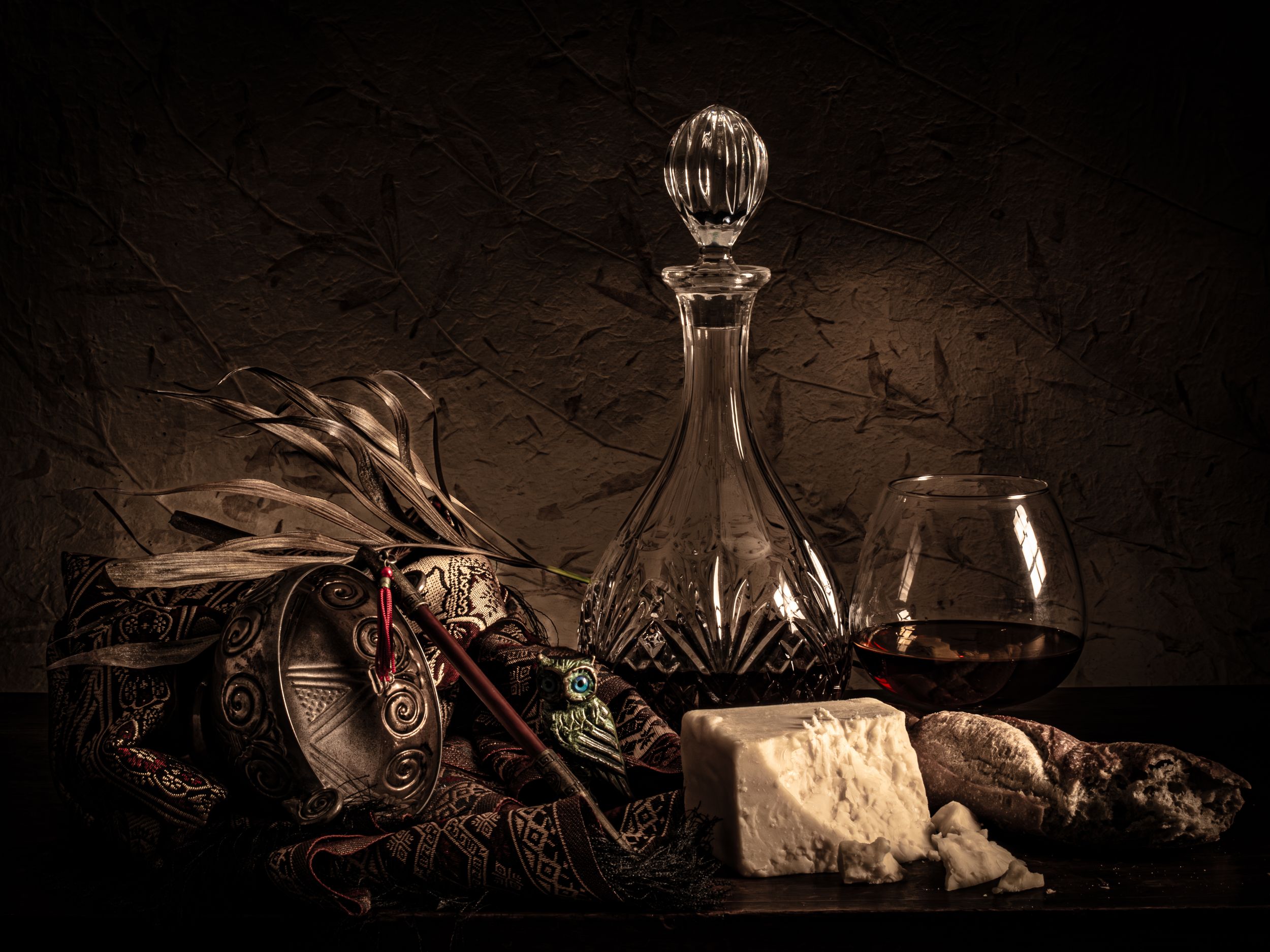 Dark still life with crystal decanter, wine glass, cheese, and ornate fabric arrangement on rustic surface.