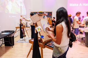 Artists painting on easels at a creative event with a 'Make A Ha' sign visible in the background.