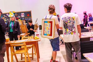 People gather at an art event, one person holding a framed 'Make Art' sign amid colorful artwork displays.