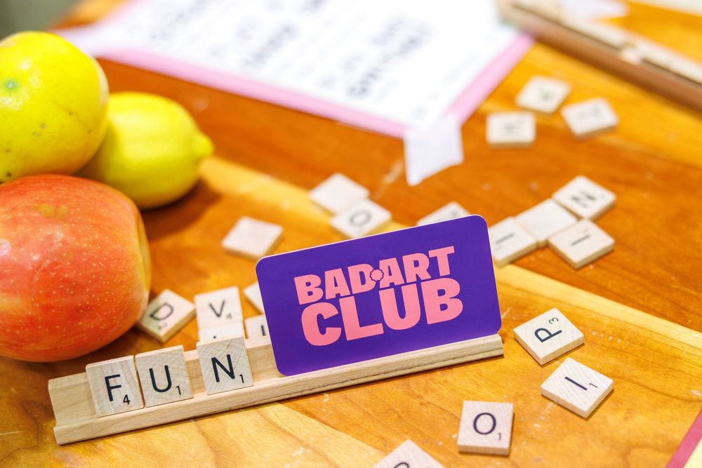 Scrabble tiles spelling FUN displayed with a Bad Art Club sign, fresh apples, and lemons on a wooden table.