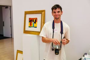 Young photographer smiling with camera at art gallery standing beside a framed colorful painting on white wall.