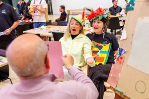 Two women wearing colorful paper hats laugh and shake hands with a man during a fun indoor craft activity event.