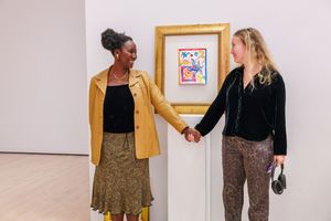 Two women holding hands and smiling beside a colorful framed artwork on a white gallery wall.