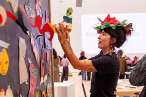 Woman wearing floral headpiece arranges colorful artwork on a gallery wall during an art exhibition event.