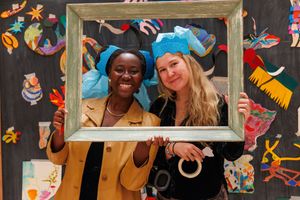 Two smiling women wearing blue paper hats hold a picture frame in front of colorful artwork.