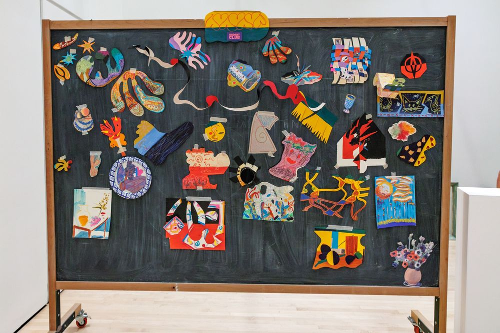 Colorful mixed-media artworks displayed on a large black chalkboard in a bright gallery.