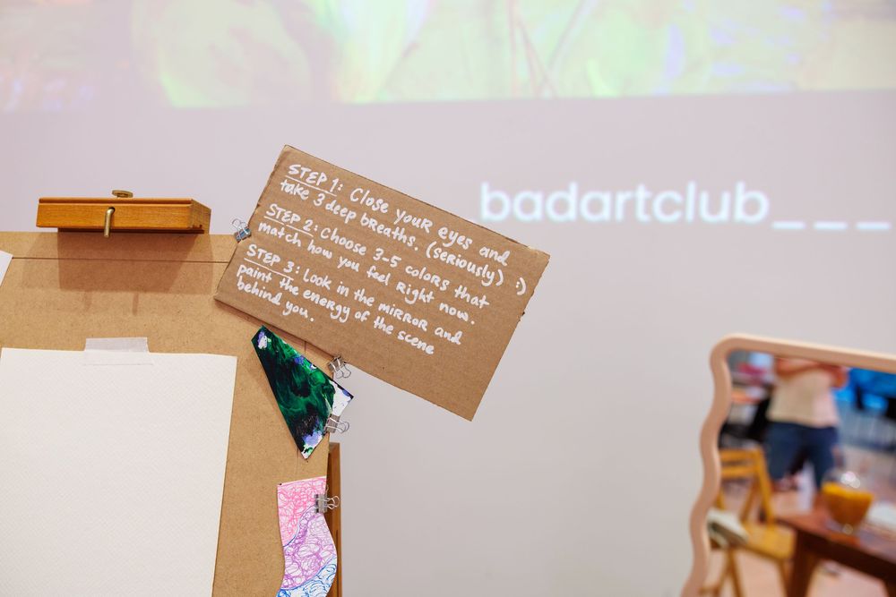 Cardboard sign displaying Bad Art Club instructions on an easel at a creative workshop event.