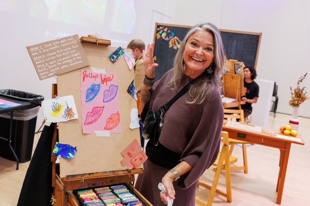 Woman smiling beside colorful art display with painted leaves at an indoor art exhibition event.