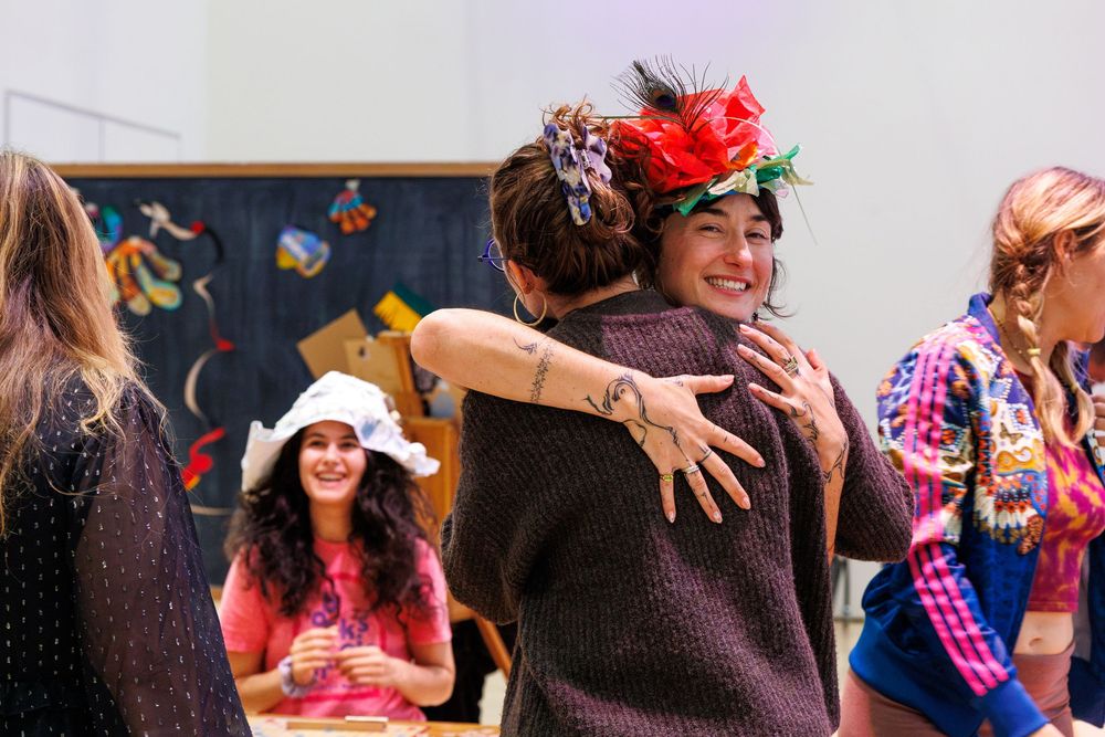 Two women hugging and smiling at a festive gathering, one wearing a colorful floral hat, with guests in background.