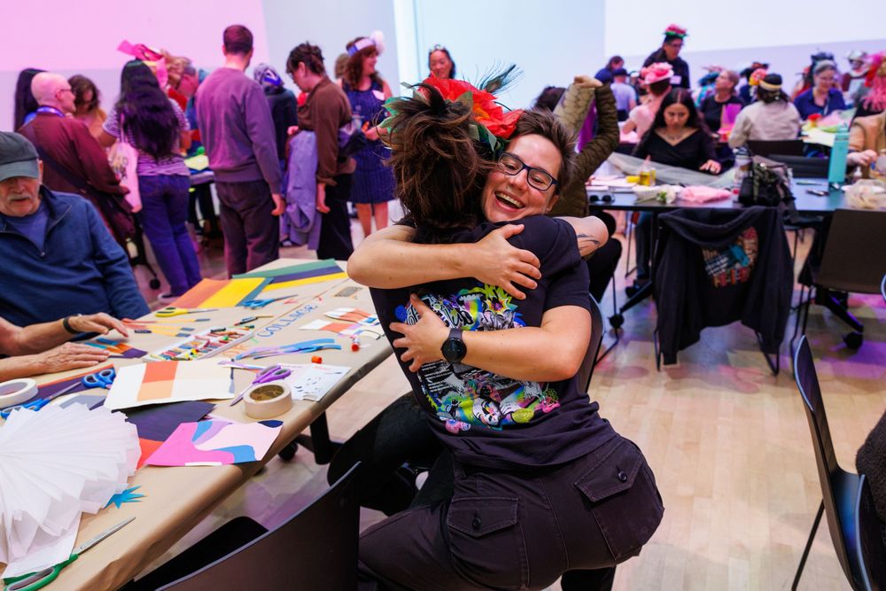 Two people share a warm hug at a colorful craft workshop event with tables and attendees in the background.