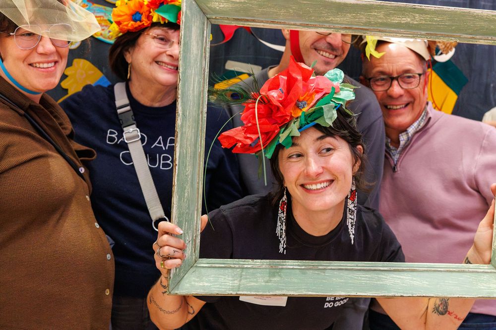 Group of five people wearing colorful floral hats pose together framing their faces with a vintage green picture frame.