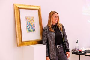 Woman smiling beside a framed floral artwork on a white pedestal in an art gallery.