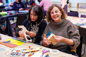 Two women smiling and crafting at a table during an art workshop, surrounded by colorful supplies.