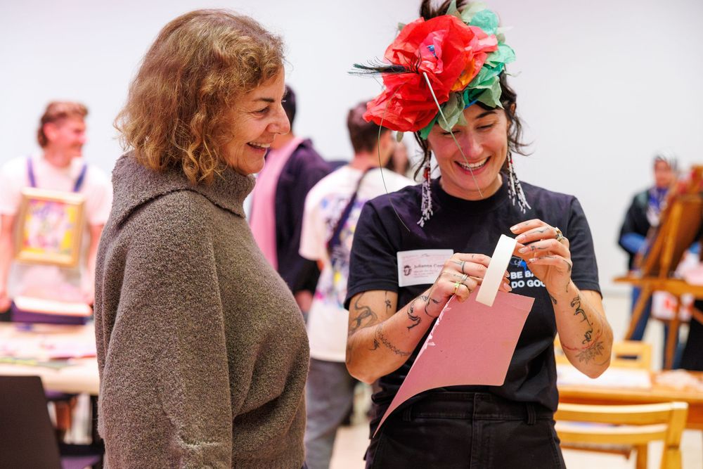 Two women smile while looking at a pink card at an indoor event, one wearing a colorful floral hat.