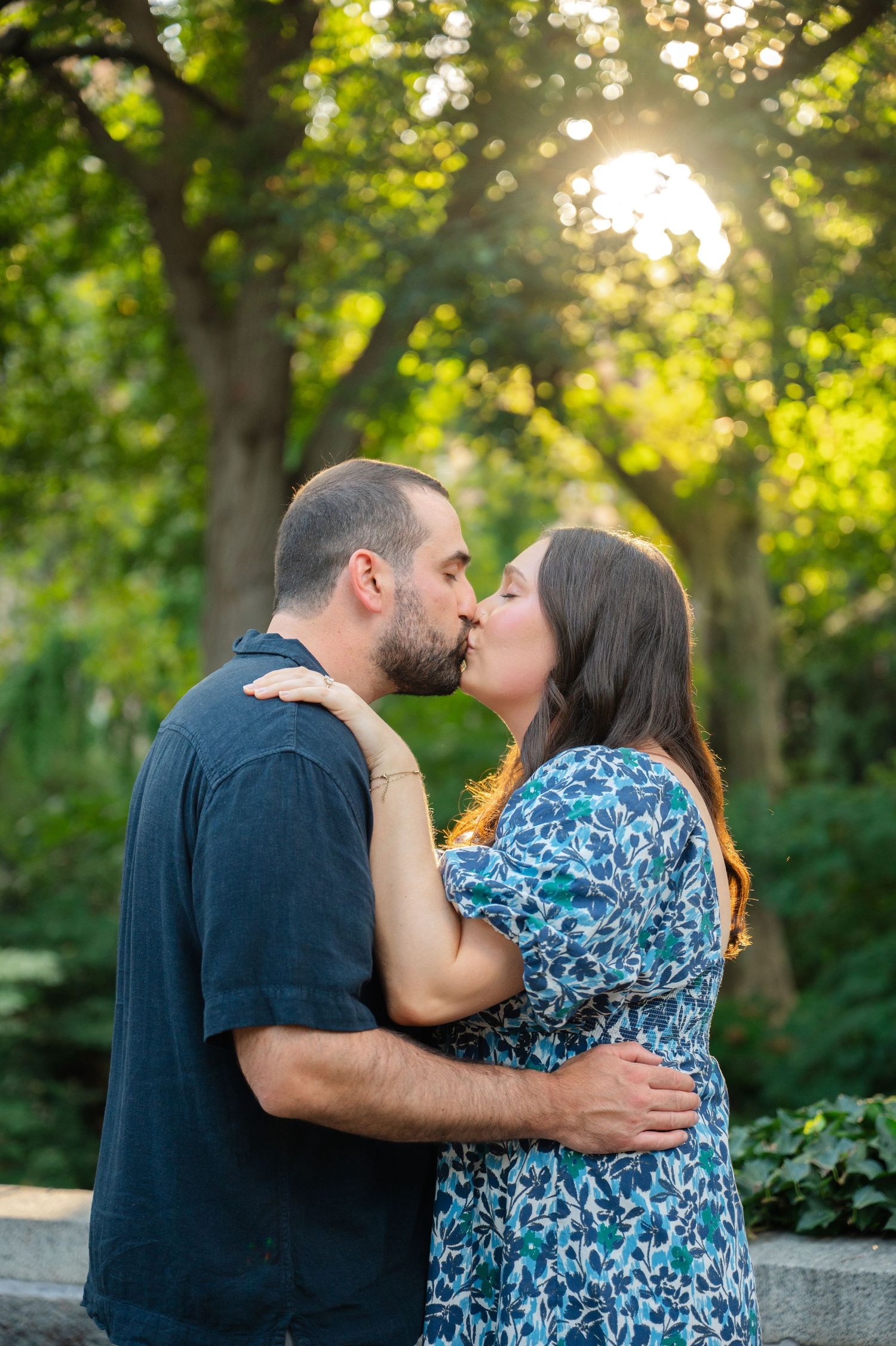 Couple sharing a romantic kiss outdoors in Carl Schurz Park in NYC, woman in floral dress embracing man in black shirt.