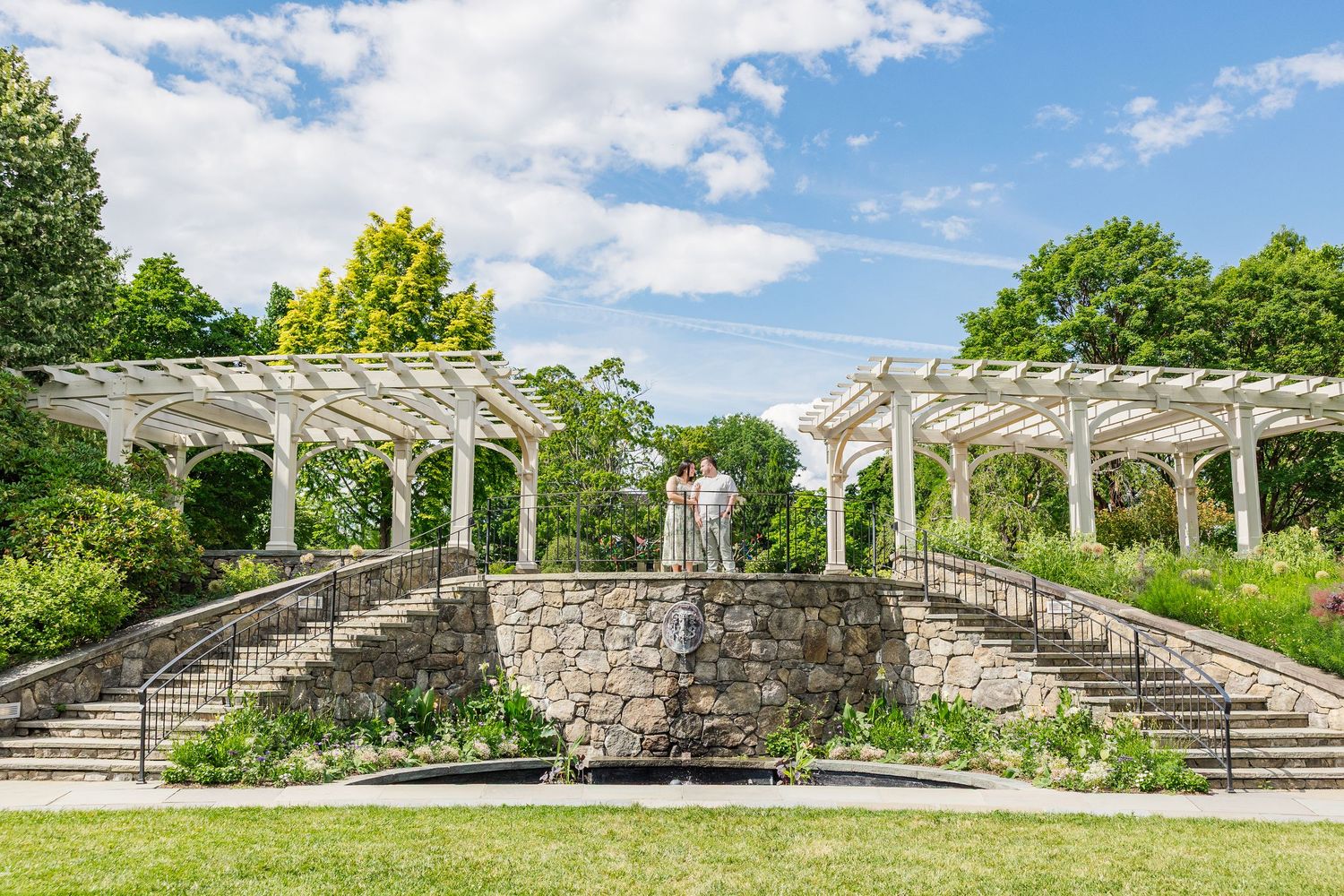 A white pergola with stone steps and a fountain in a lush green garden under a partly cloudy blue sky.