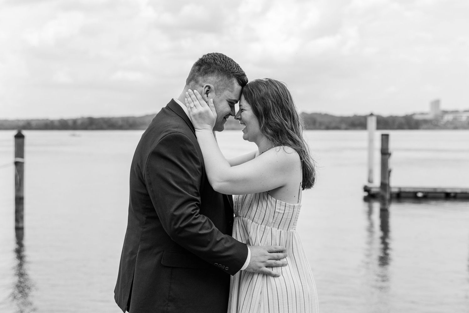 Black and white photo of a couple sharing a romantic forehead touch near a waterfront dock.