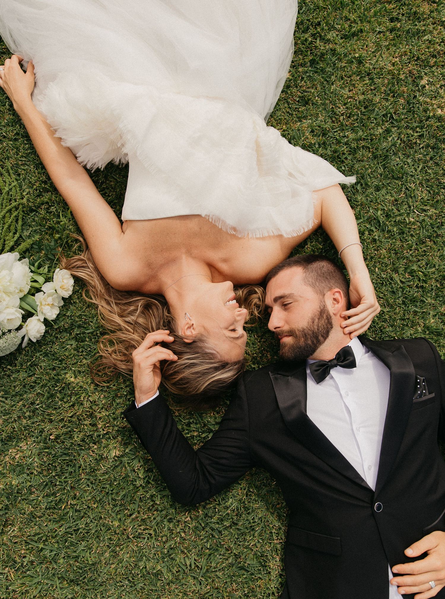 Bride and groom lying on green grass, smiling at each other, wearing wedding dress and black tuxedo with bow tie.