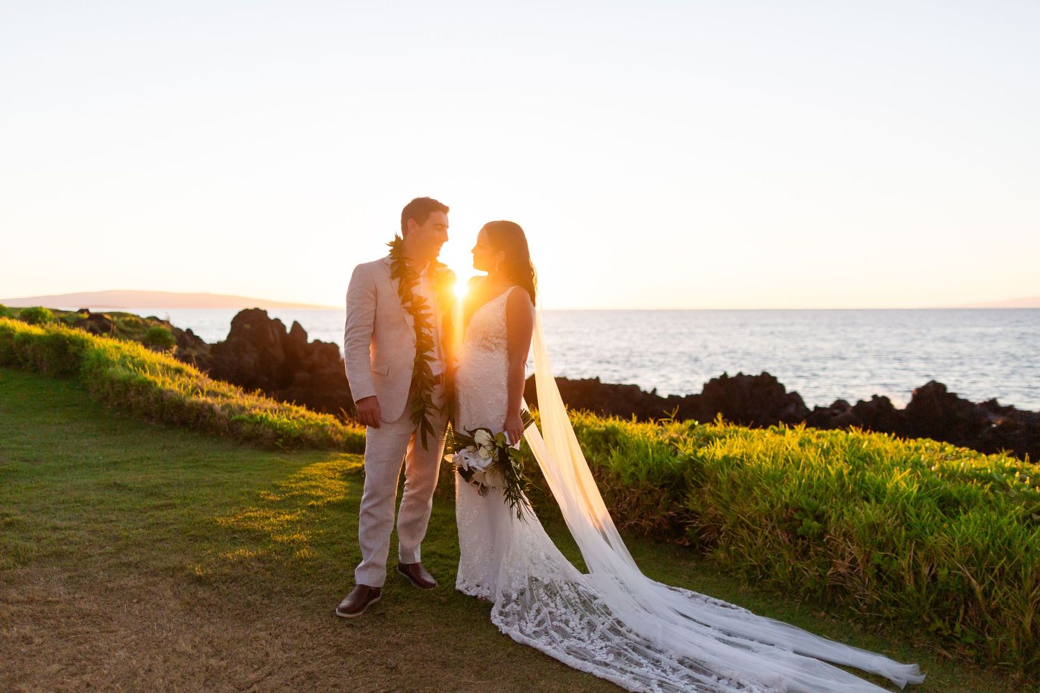 Bride and groom share a romantic sunset moment on a coastal cliff in Maui, with ocean views and golden light.