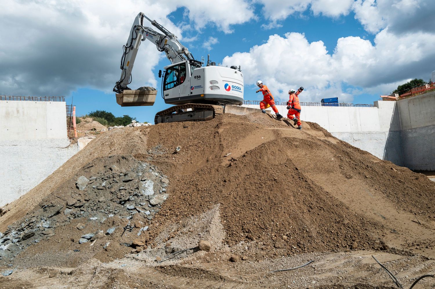 Construction workers in orange safety gear walk beside a white excavator on a large dirt mound at an active building site.