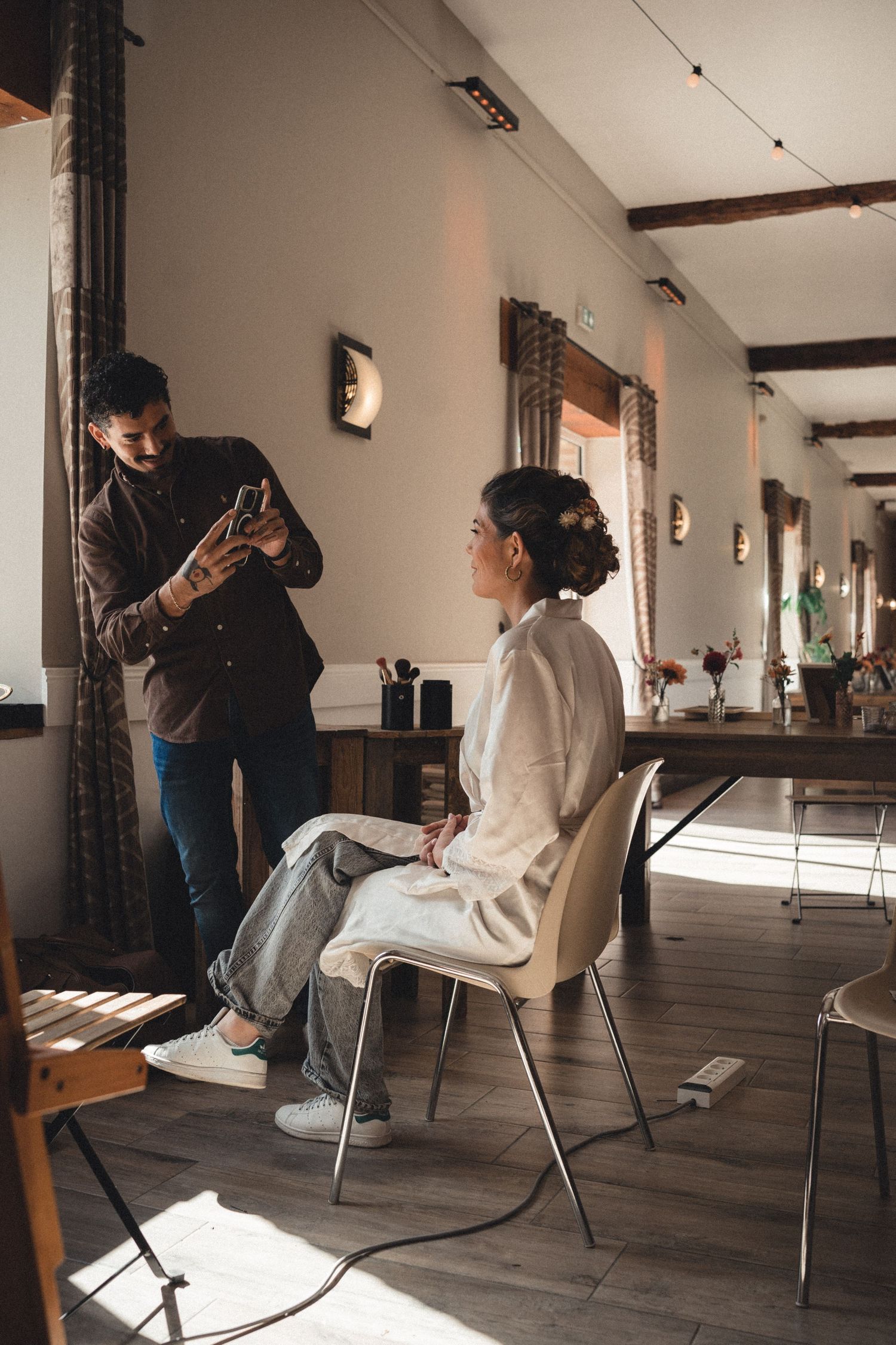 A bride sits in a chair while a photographer captures her look in a sunlit rustic venue before the wedding ceremony.