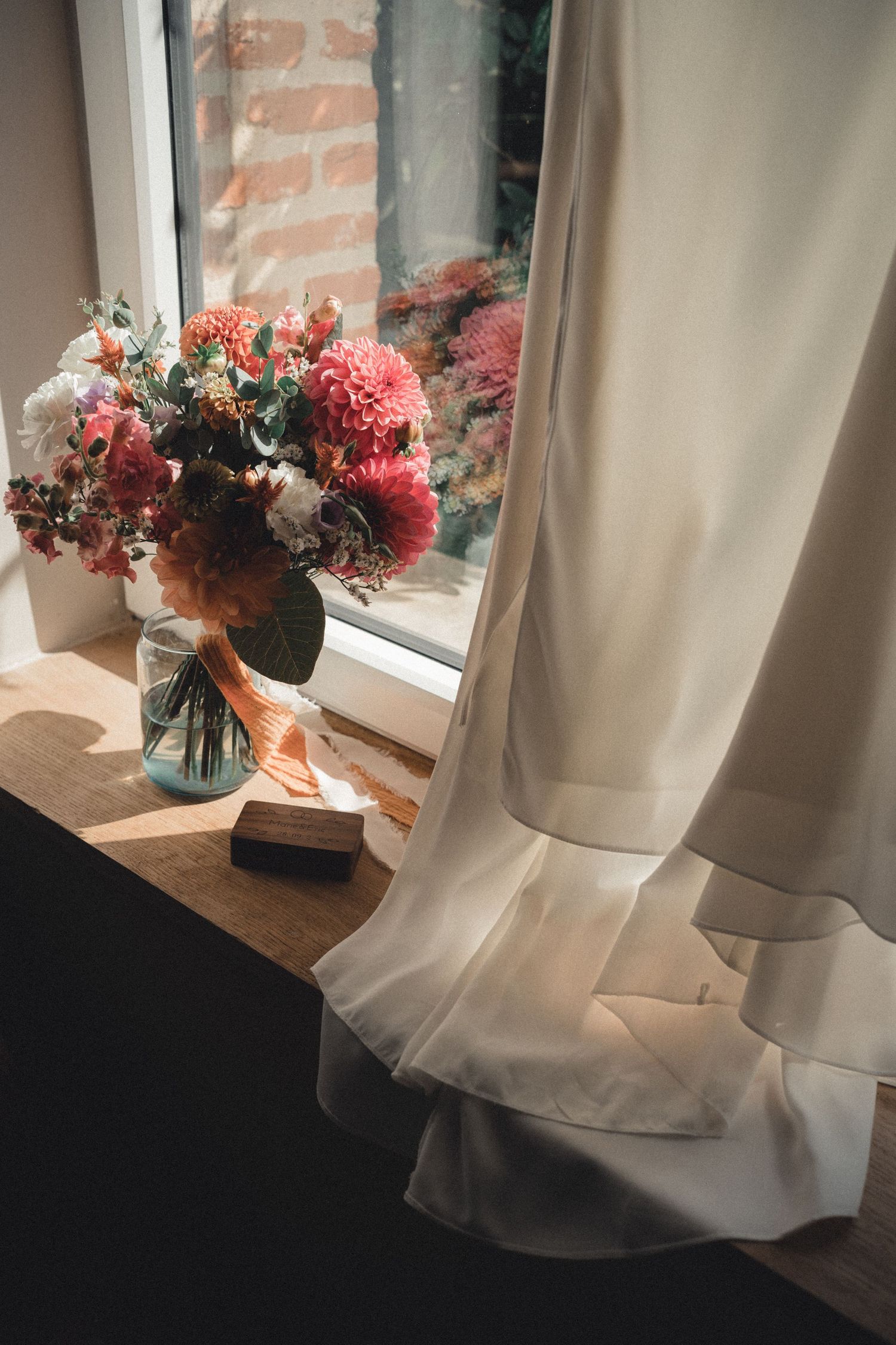 Bridal bouquet of pink and white flowers in a glass vase on a windowsill beside a white wedding dress.
