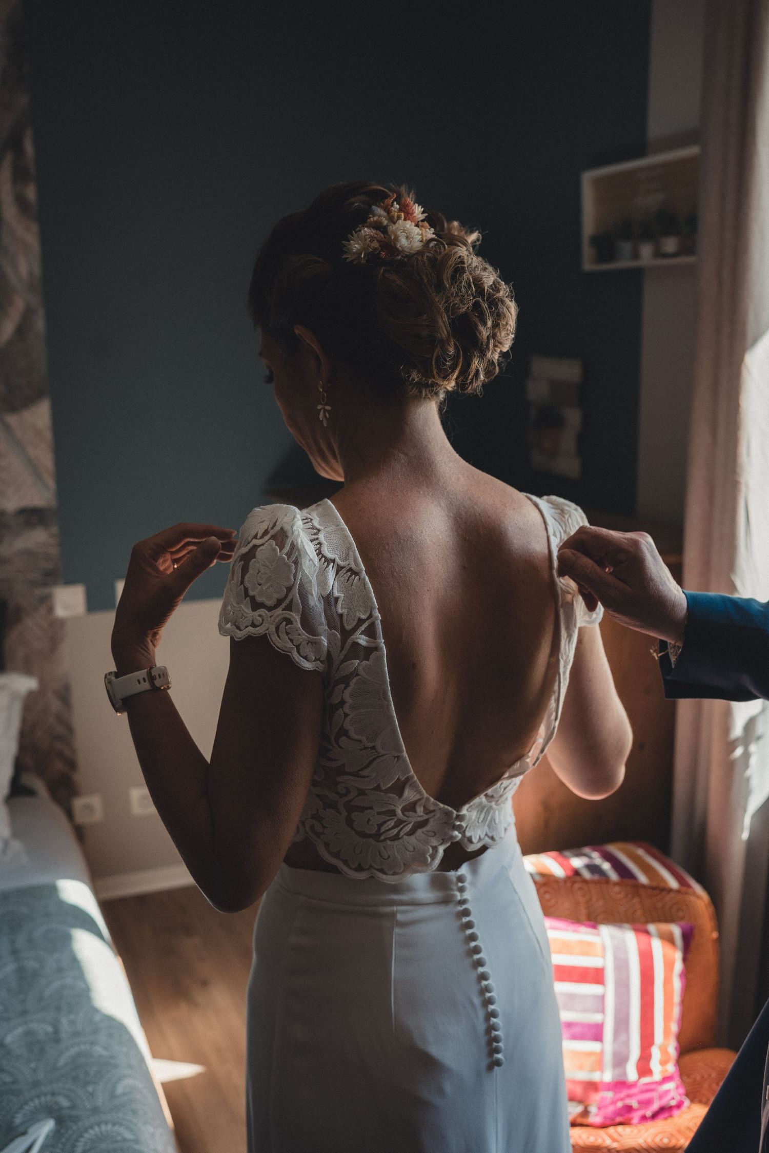 Bride getting her lace open-back wedding dress fastened by a helper in a sunlit bedroom.