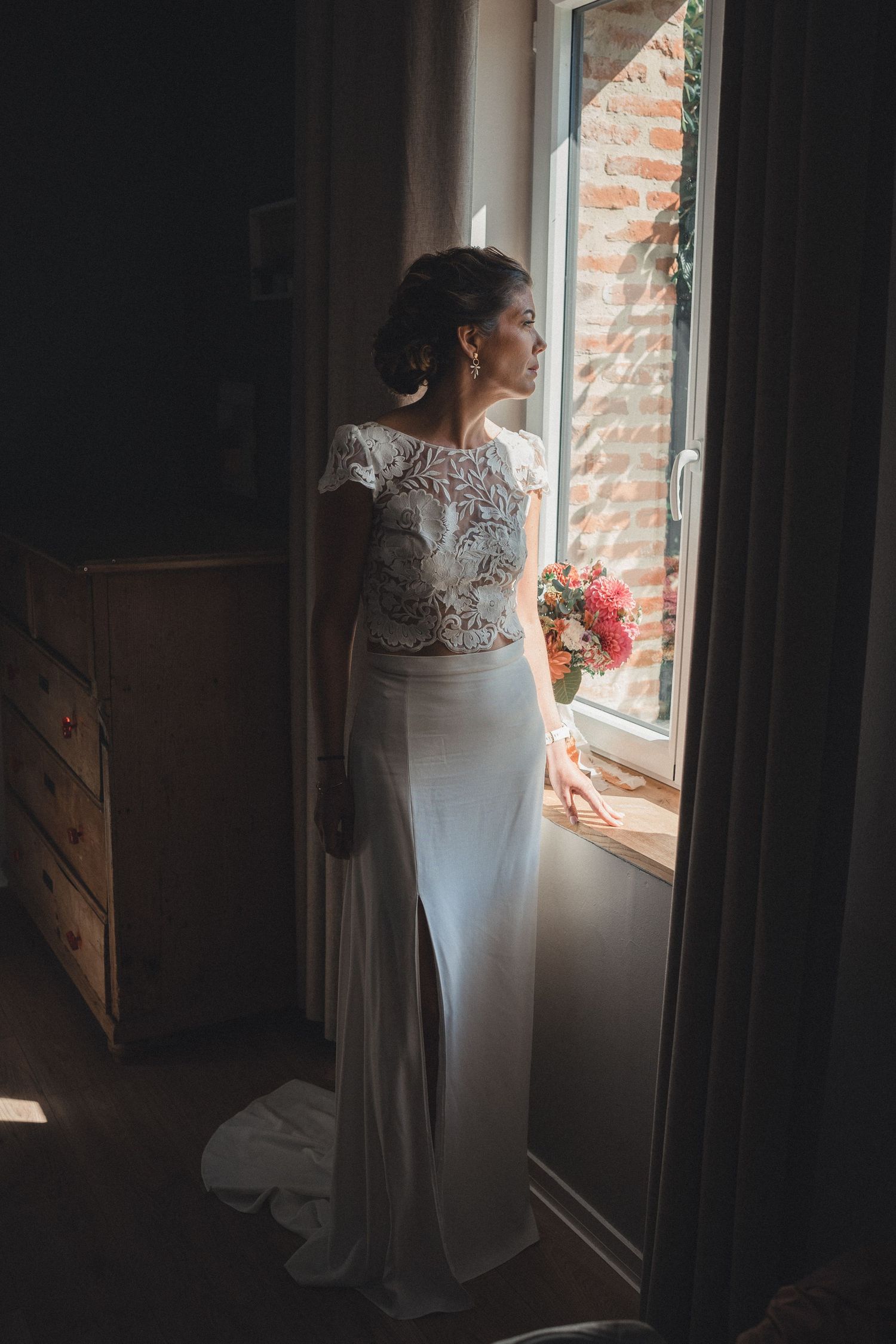 Bride in a white lace wedding dress holding a bouquet while gazing out a sunlit window.