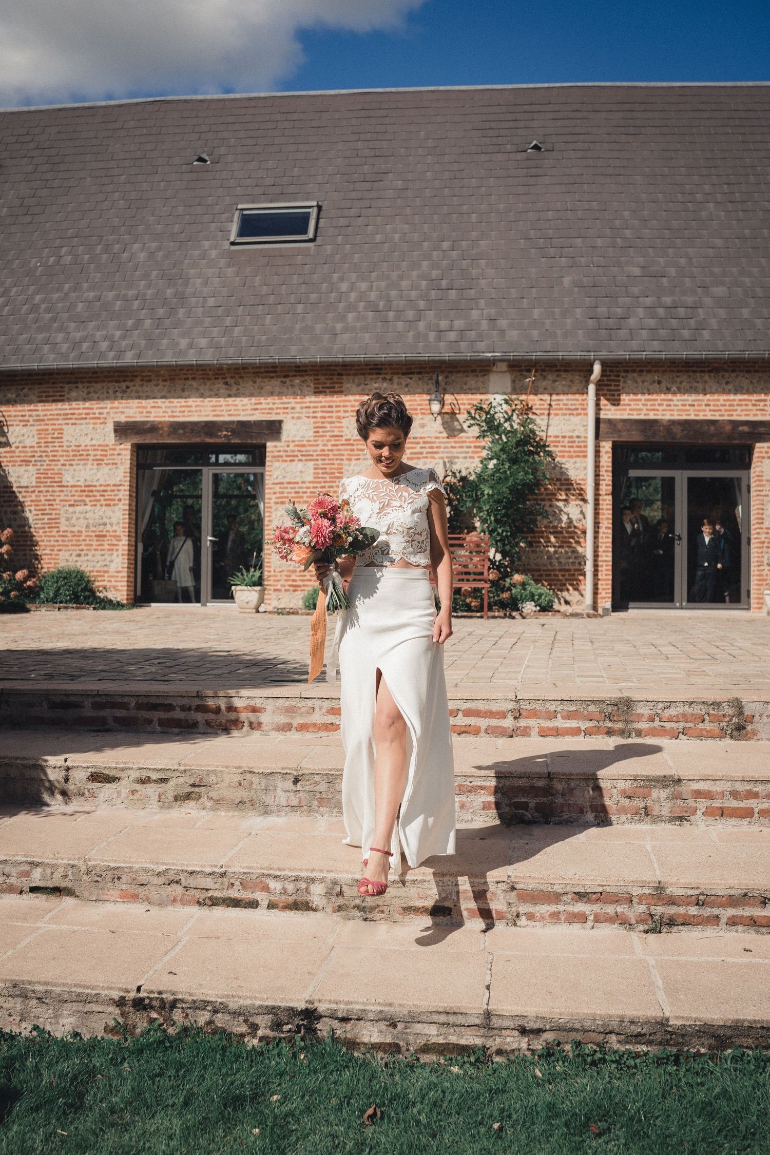 Bride in white lace and satin wedding dress with slit holding pink bouquet on brick venue steps.