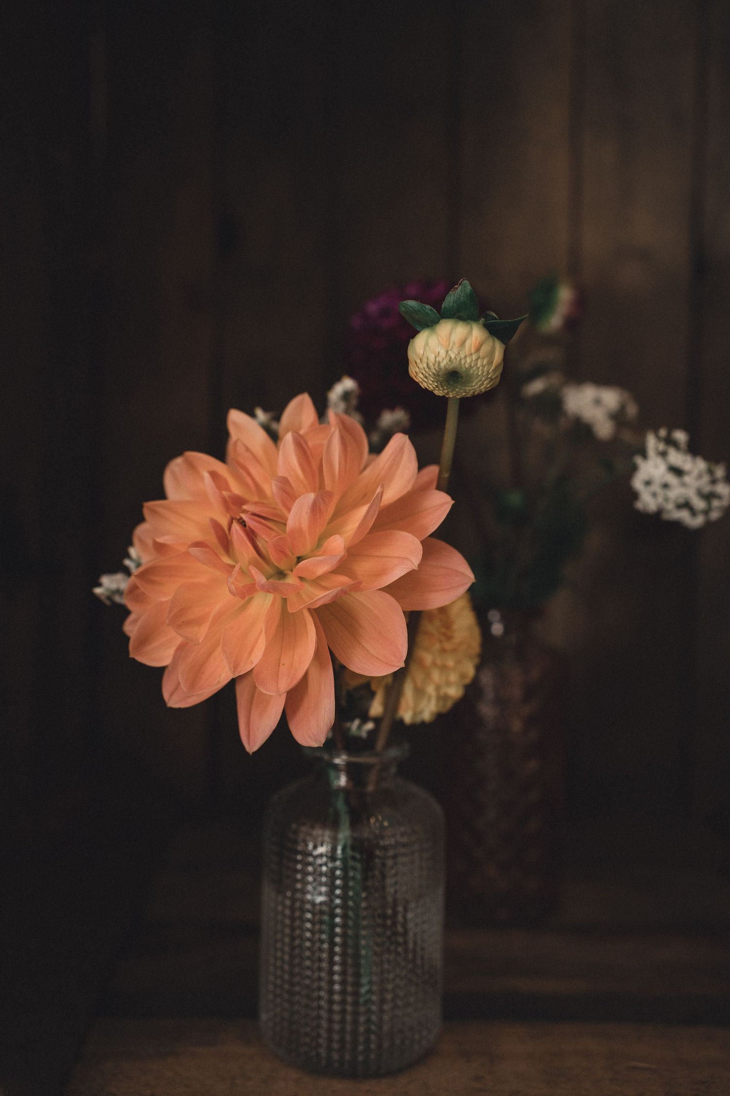 Peach dahlia and mixed wildflowers in a glass vase against a dark rustic wooden background.