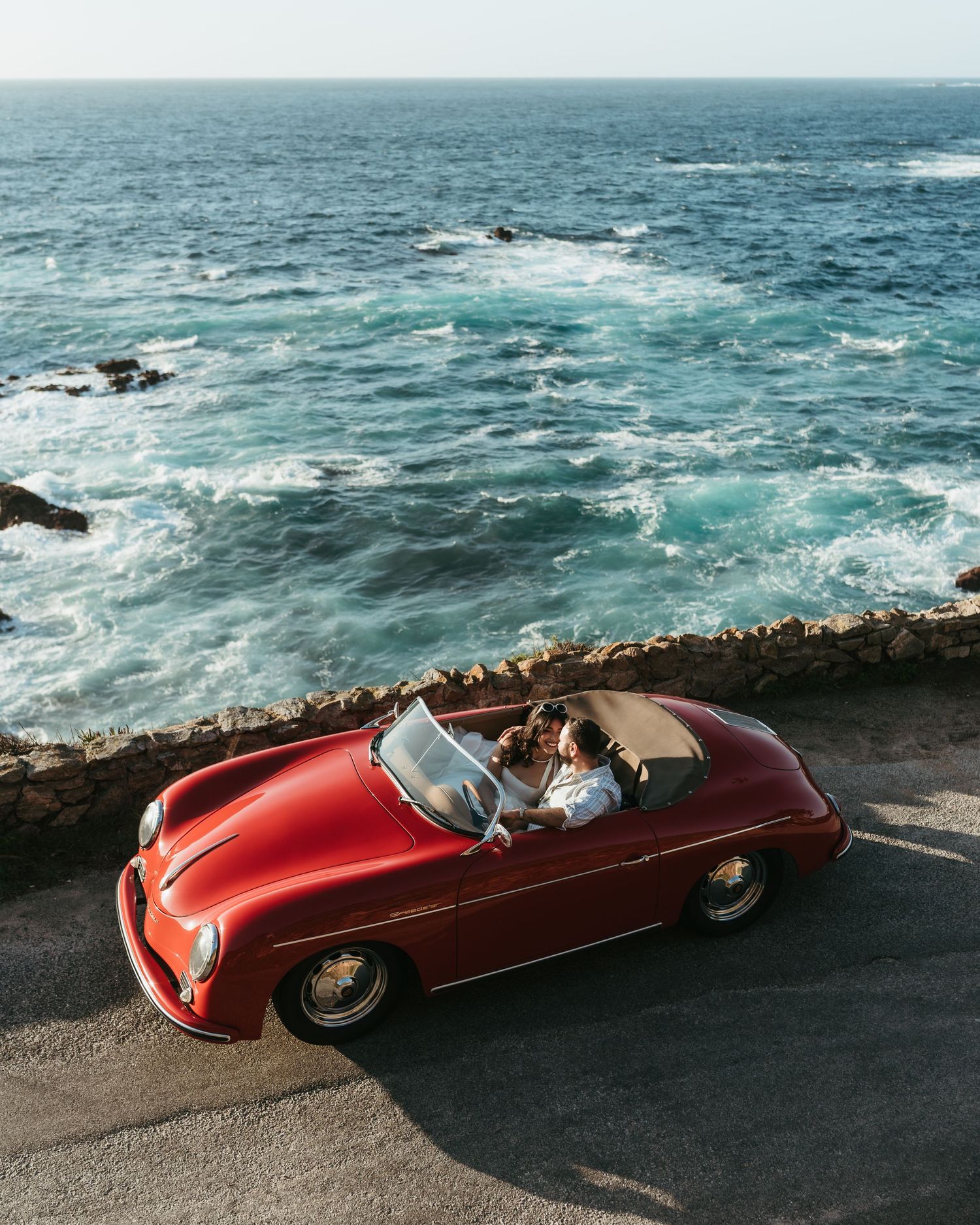 Red vintage Porsche 356 convertible parked on coastal road with turquoise ocean waves in background.