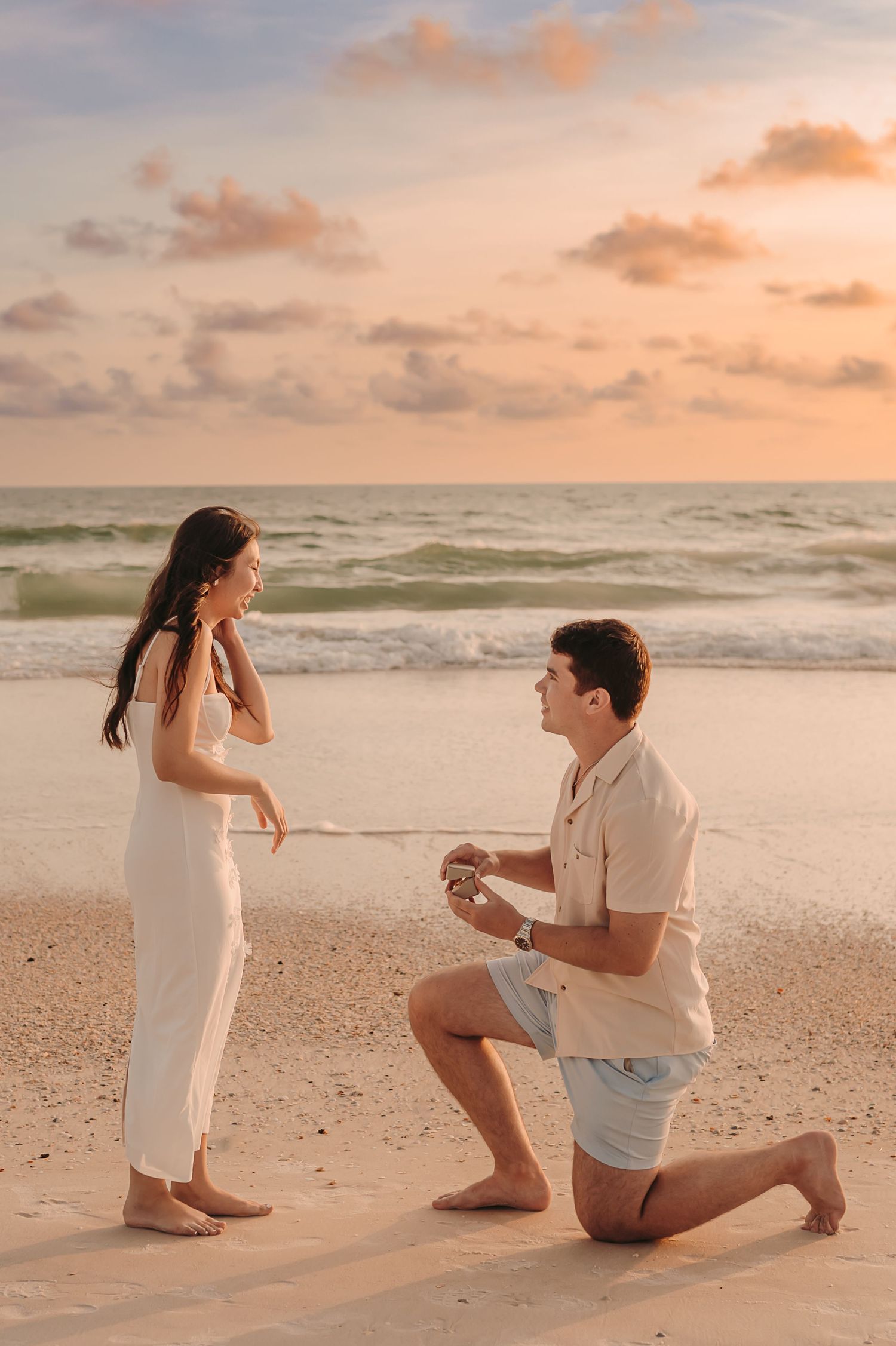 Man proposes on bended knee to woman in white dress on a sandy beach at sunset.