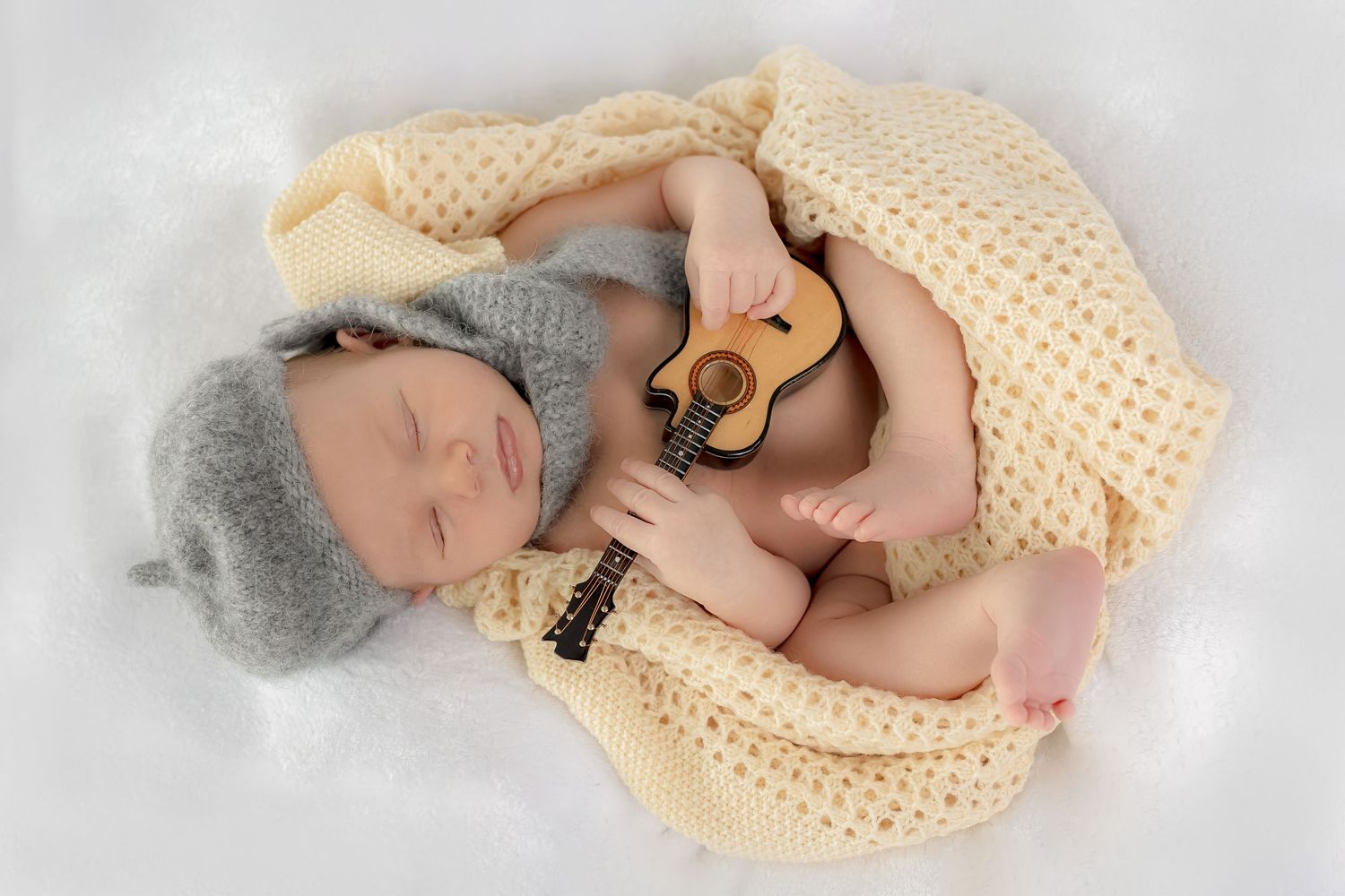 Sleeping newborn baby in gray knit hat holds tiny guitar wrapped in yellow knit blanket.