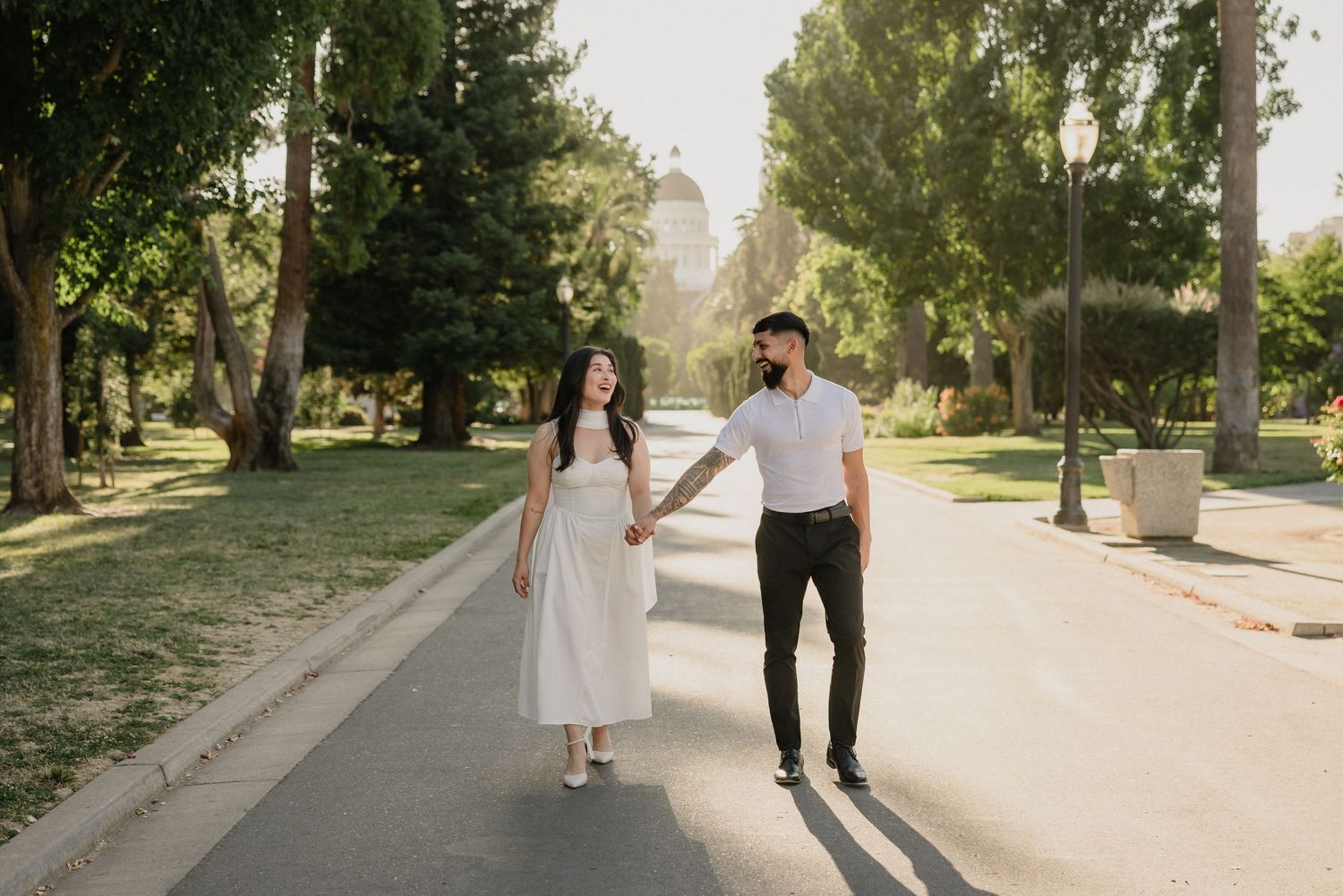 Couple holding hands walking on a sunny park path, woman in white dress, man in white shirt and black pants.