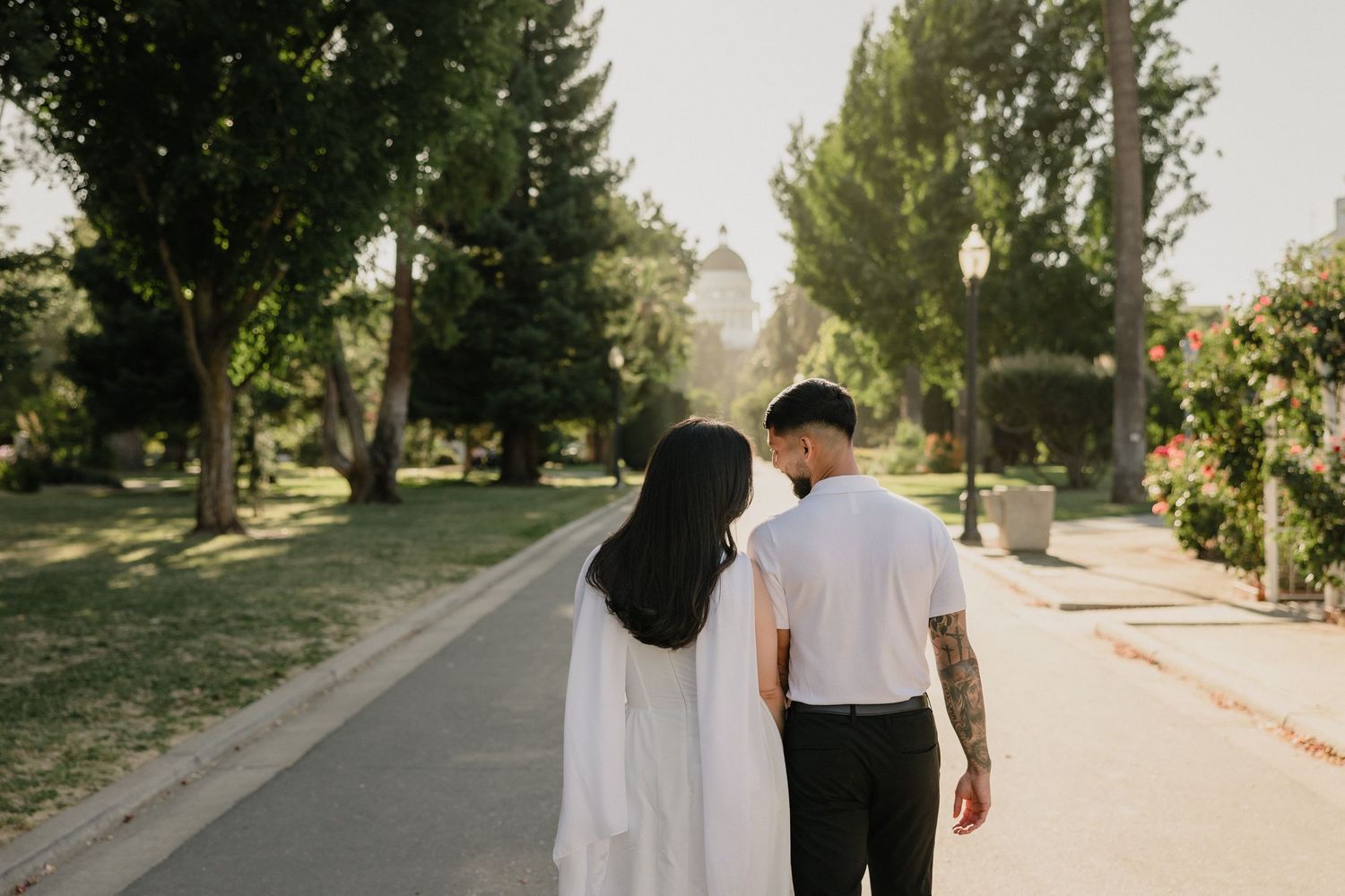 Couple walking hand in hand through a sunlit park path, man with tattoos and woman in white dress kissing.