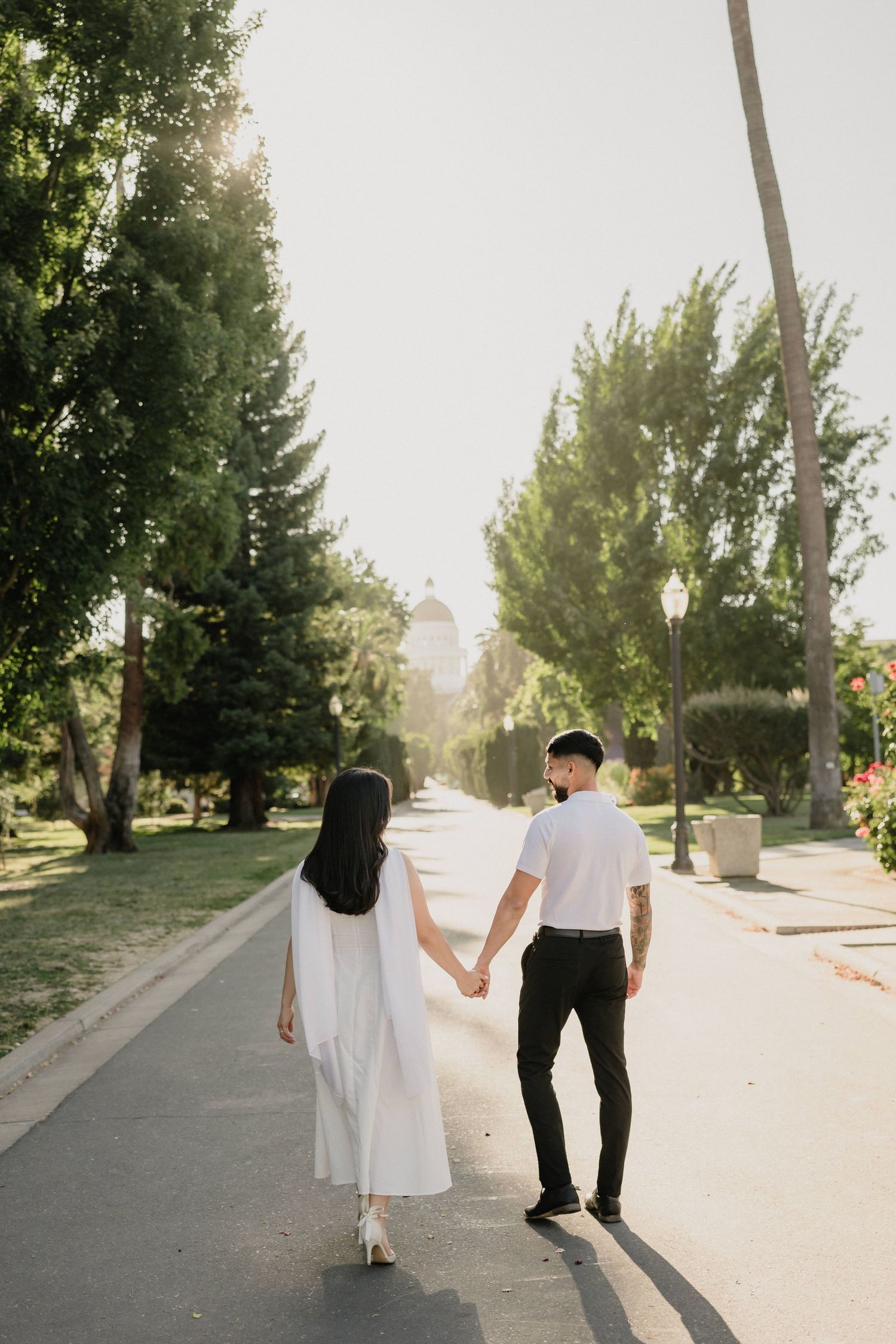 Couple holding hands walking down a sunny tree-lined path, woman in white dress, man in white shirt and black pants.