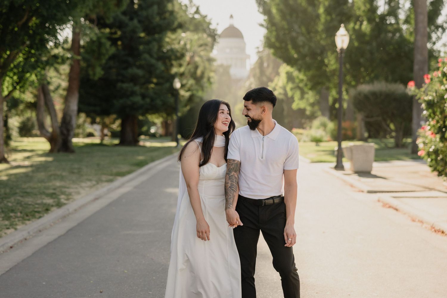 Romantic couple gazing at each other on a sunlit park path, woman in white dress, man with tattoos.