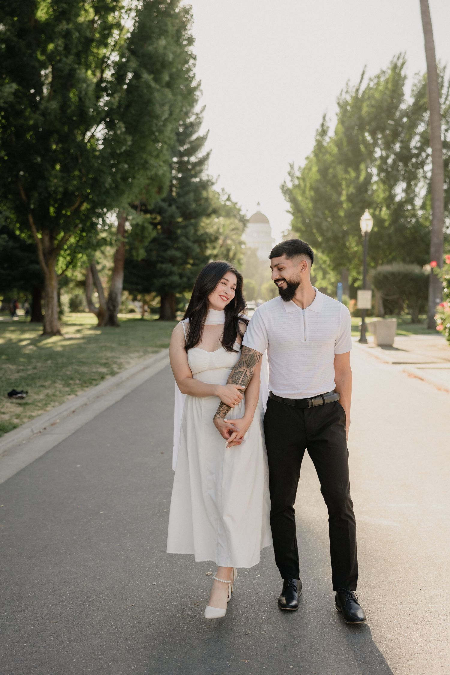 Couple holding hands walking on a tree-lined path, woman in white dress and man in white shirt and black pants.