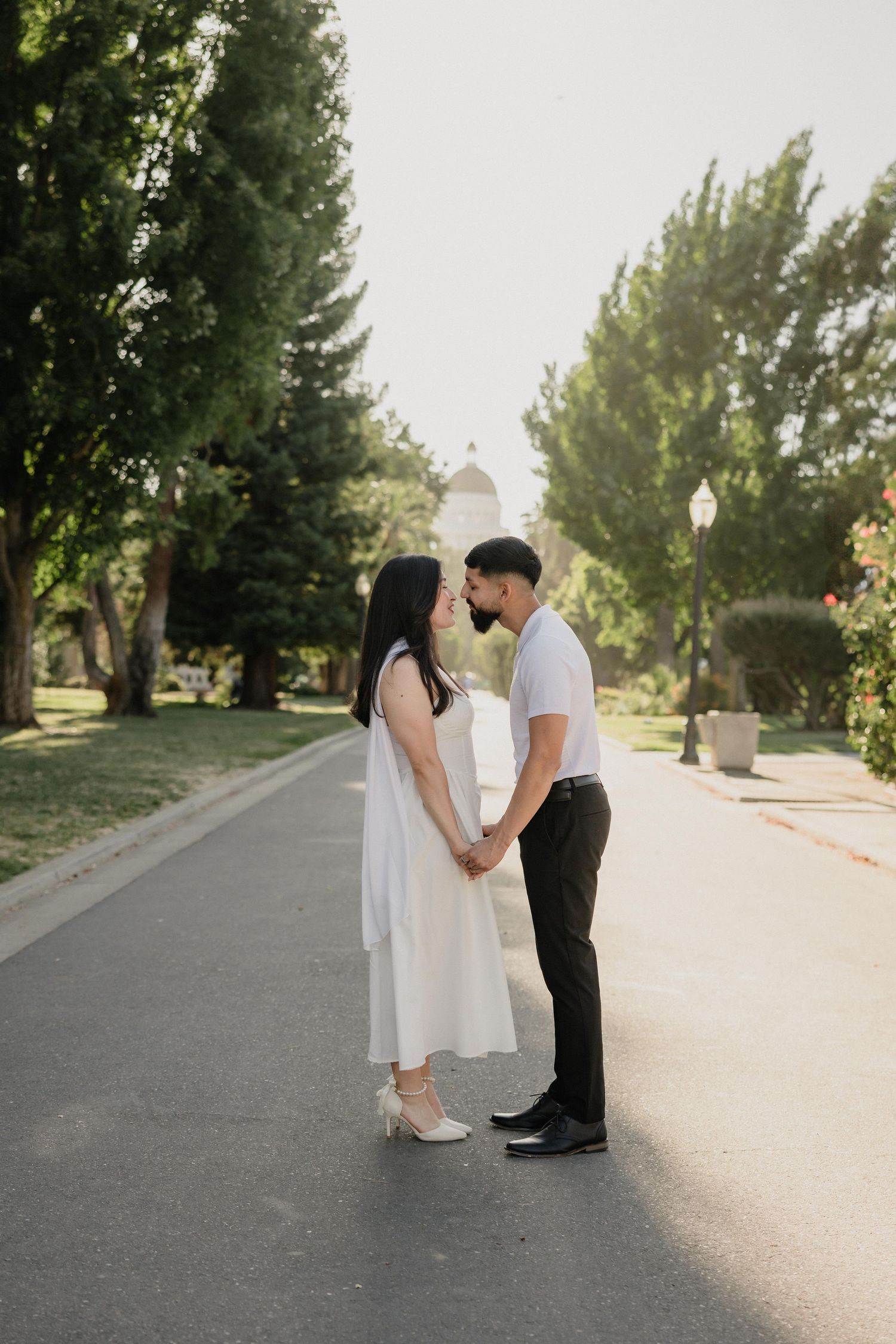 Couple holding hands and sharing a kiss on a tree-lined path in a sunny park, woman in white dress.